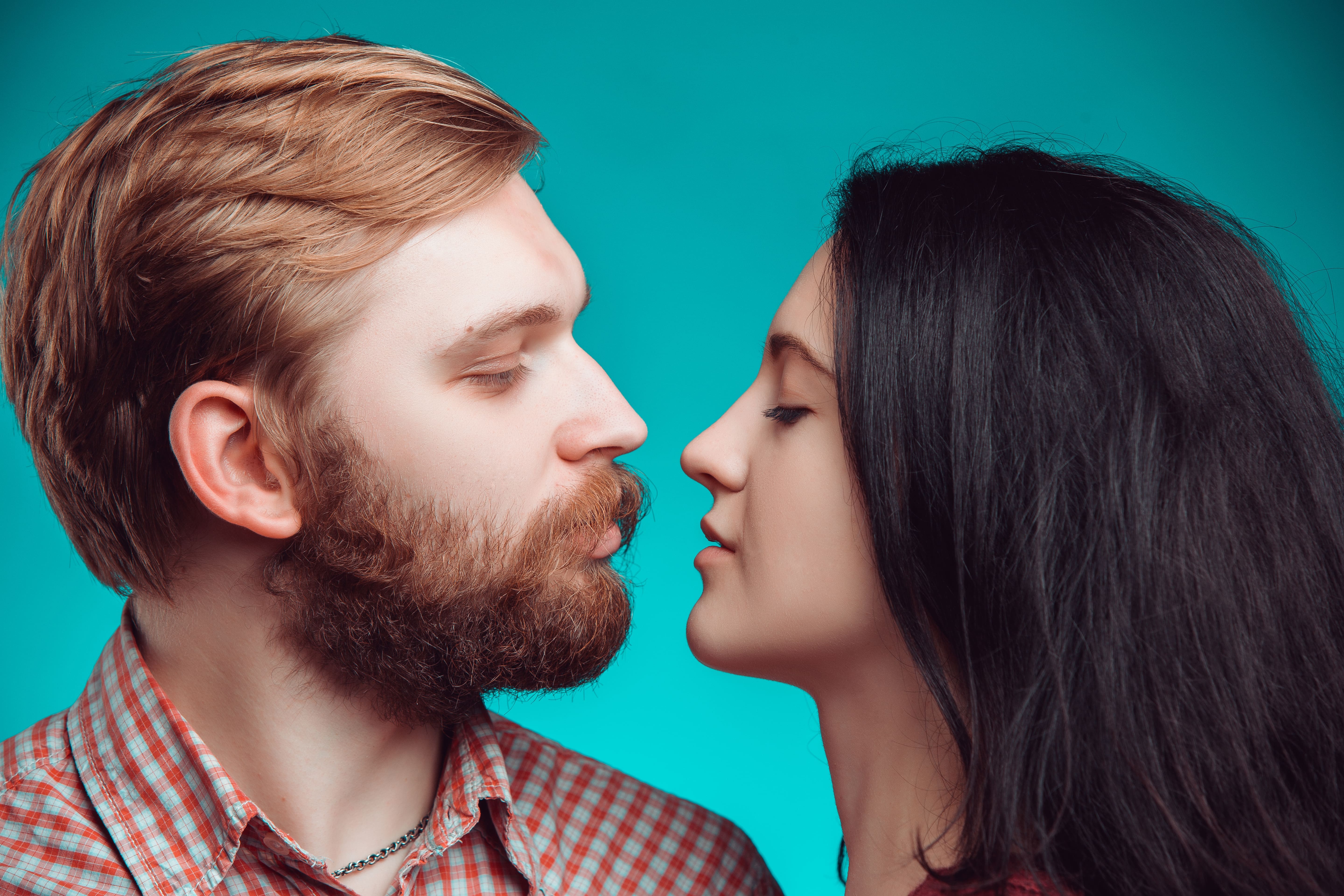 Close-up of a man and woman facing each other, noses touching, on the cusp of a kiss, set against a teal background.